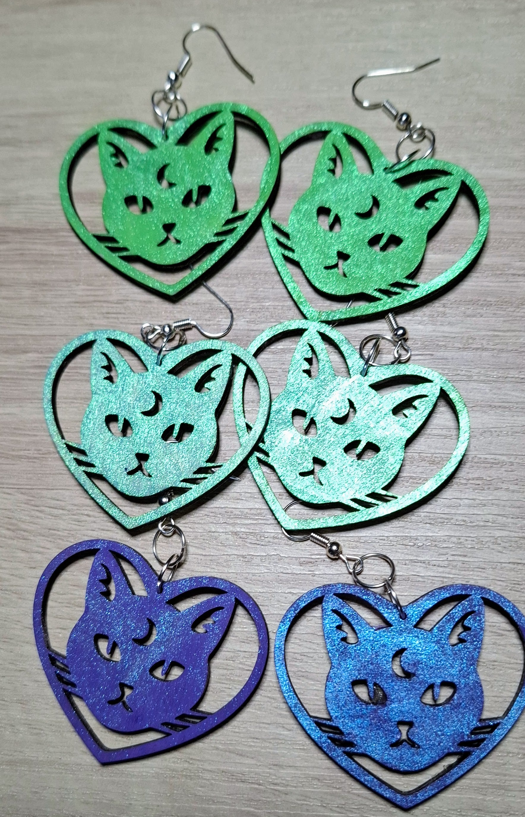 Green, teal, and purple cat-shaped heart earrings on a wooden surface.