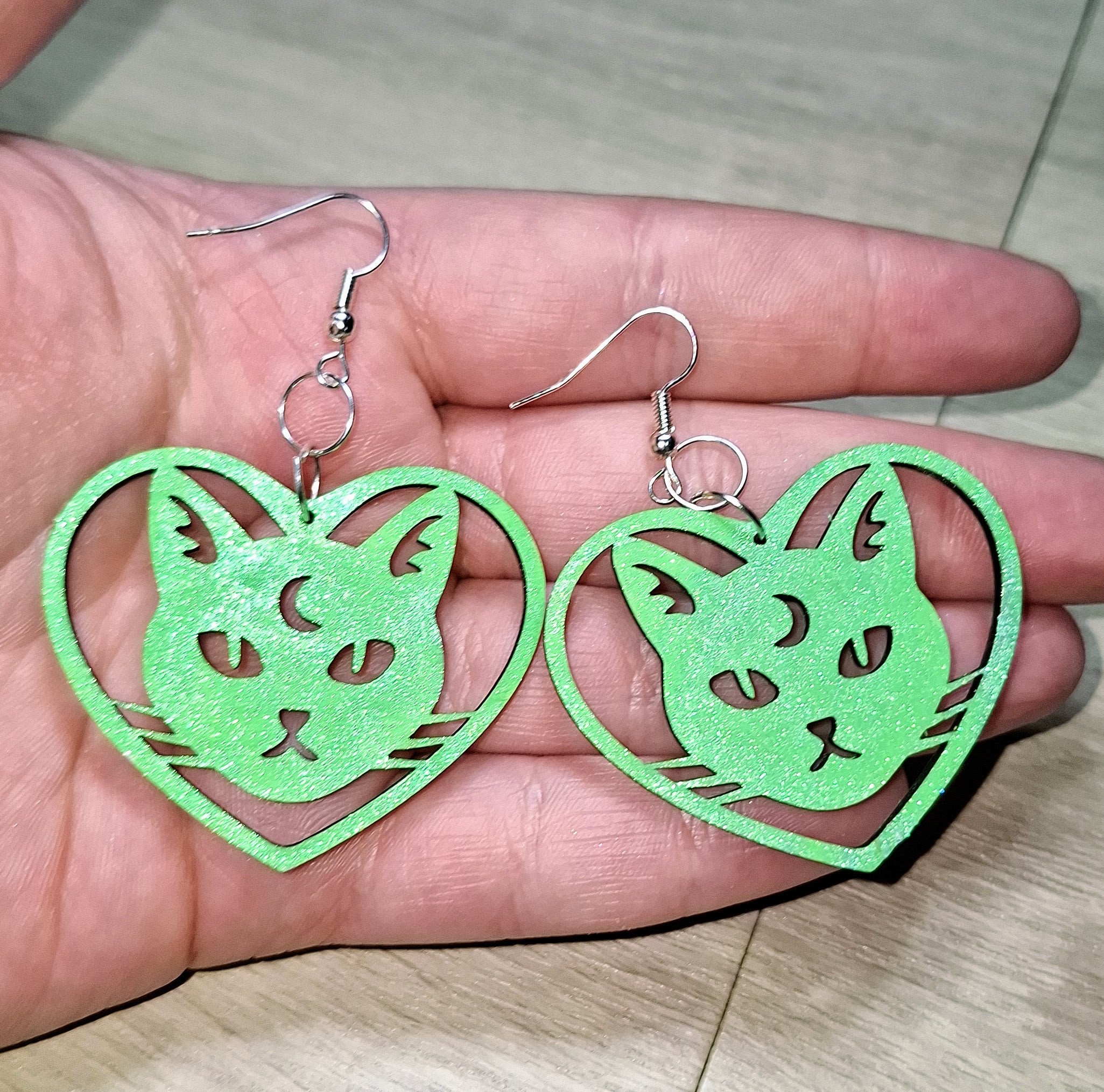 Green cat-shaped heart earrings held in a hand on a light wooden surface.