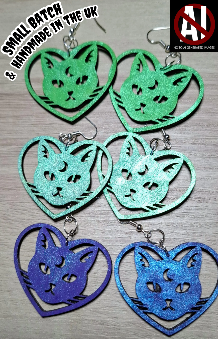 Green, teal, and purple cat-shaped heart earrings on a wooden surface.