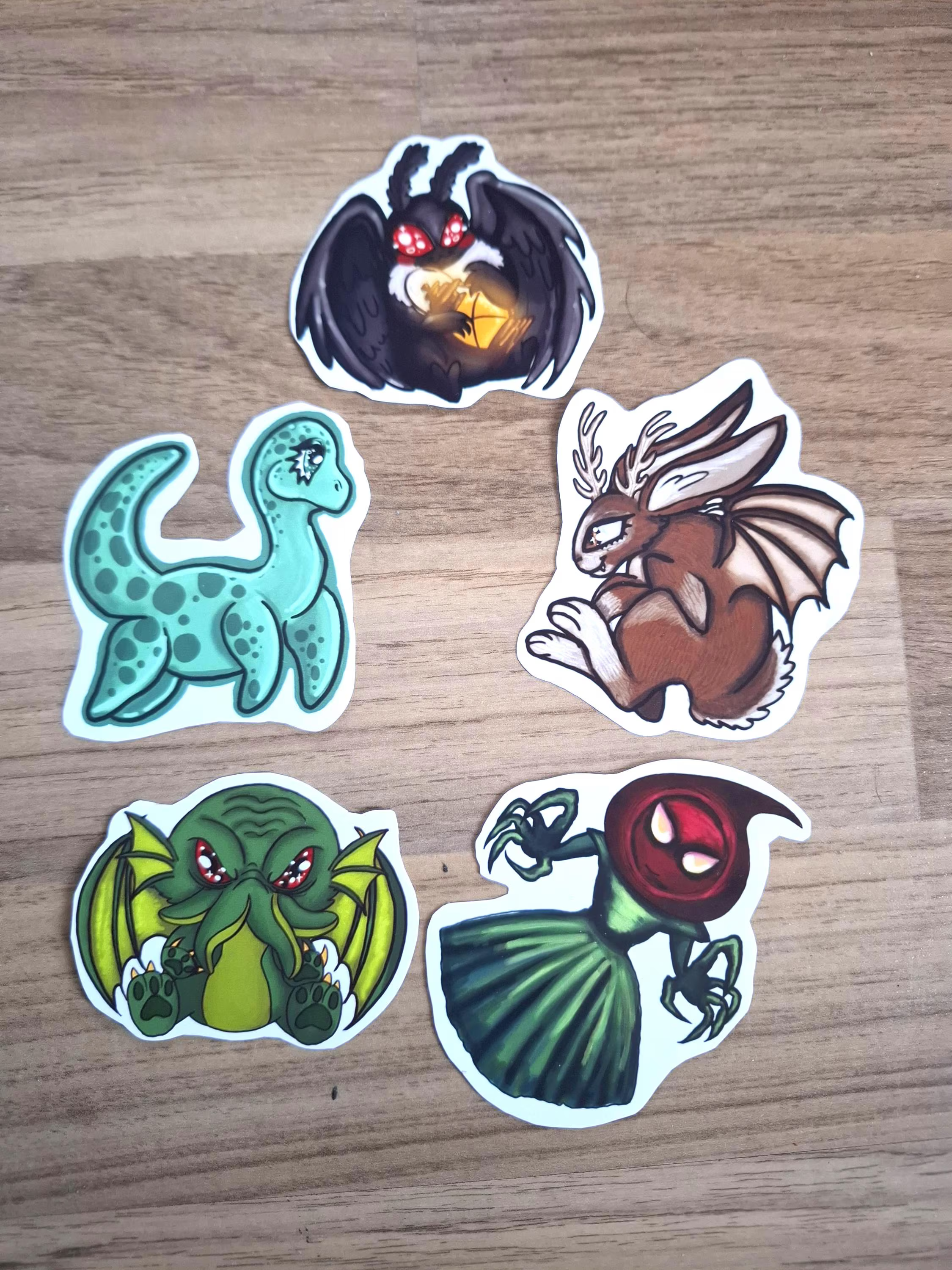Five colorful stickers of fantasy cryptid  creatures on a wooden surface