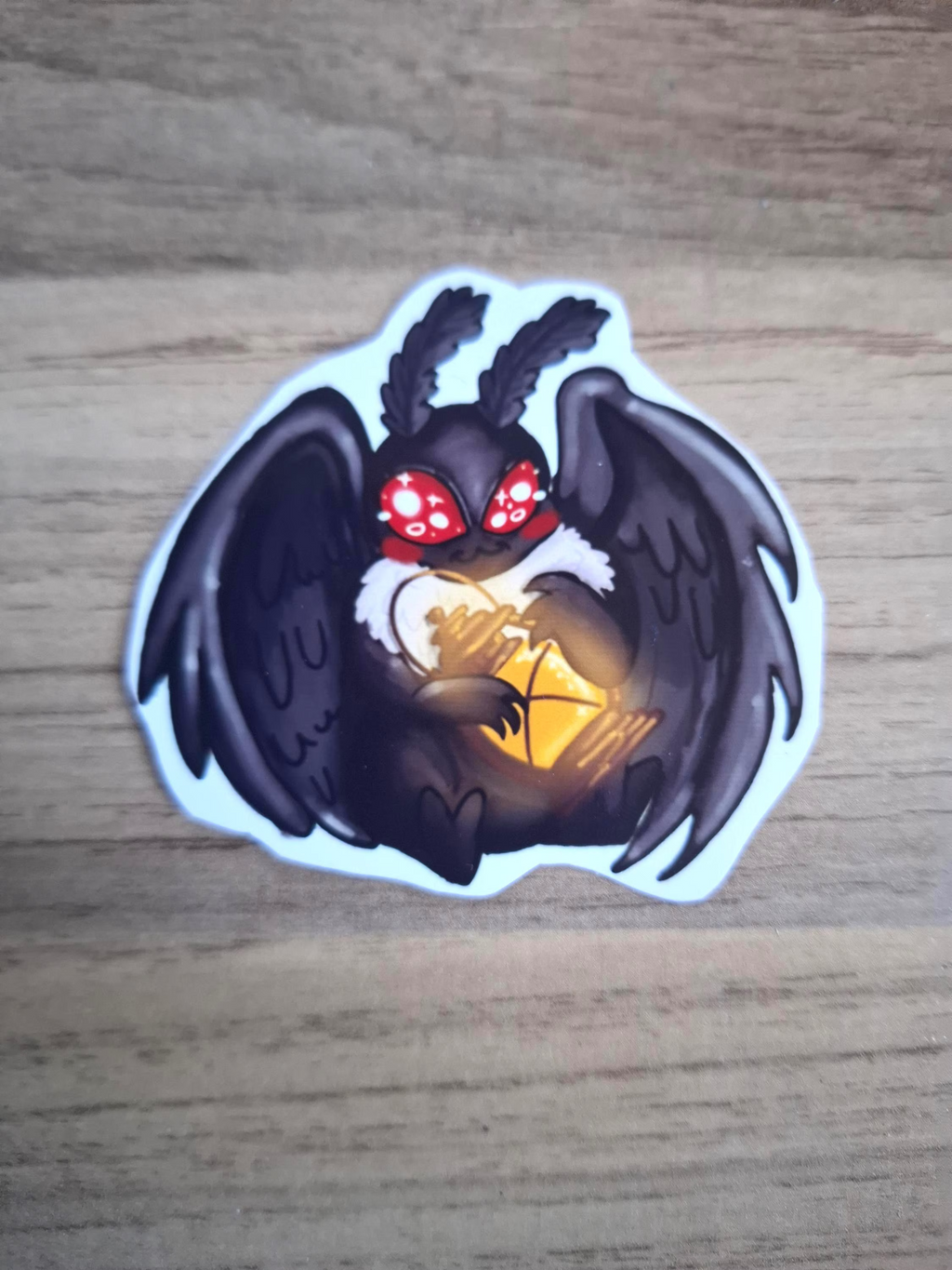 Sticker of a dark moth creature with red eyes and glowing yellow center on a wooden surface