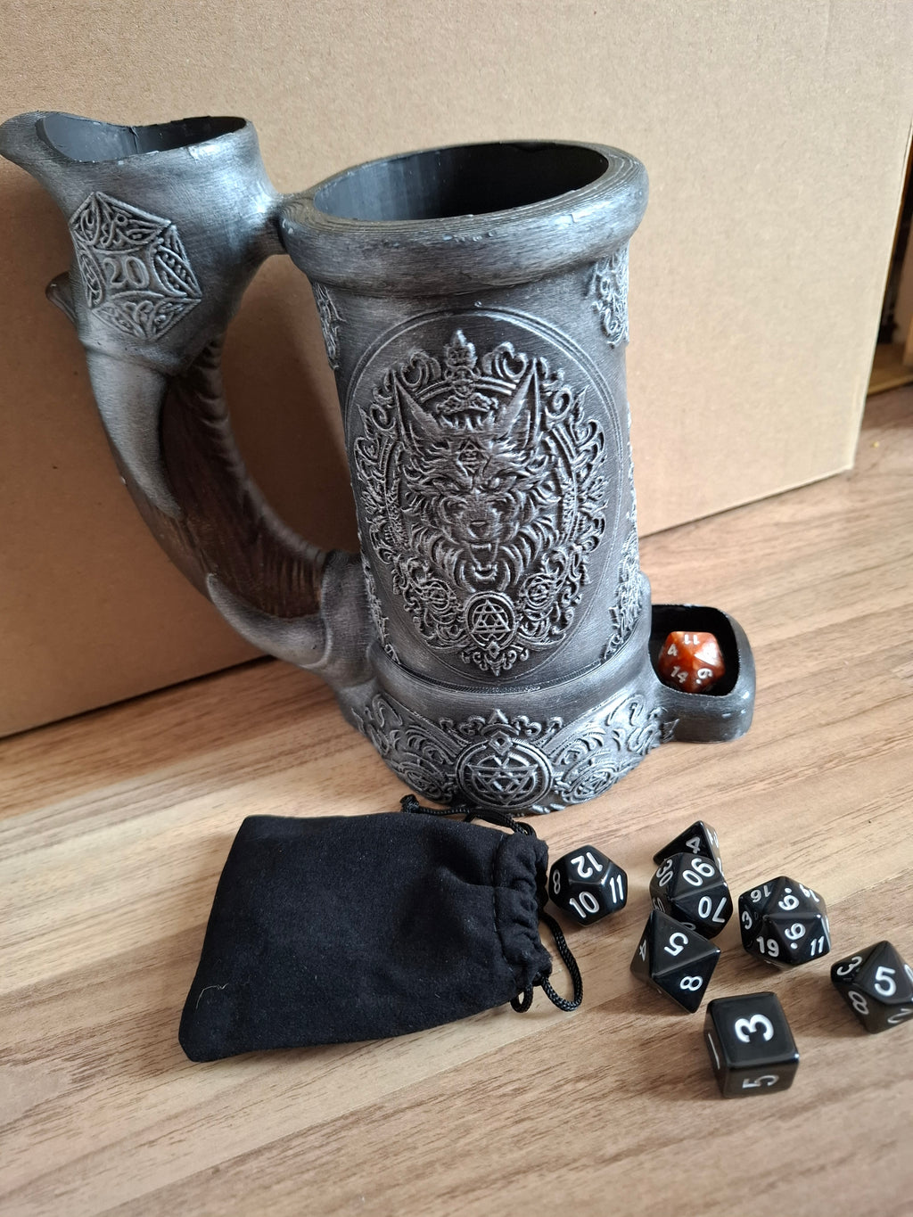 Decorative  dice holder with ornate designs on a wooden surface, surrounded by dice and a black drawstring bag.